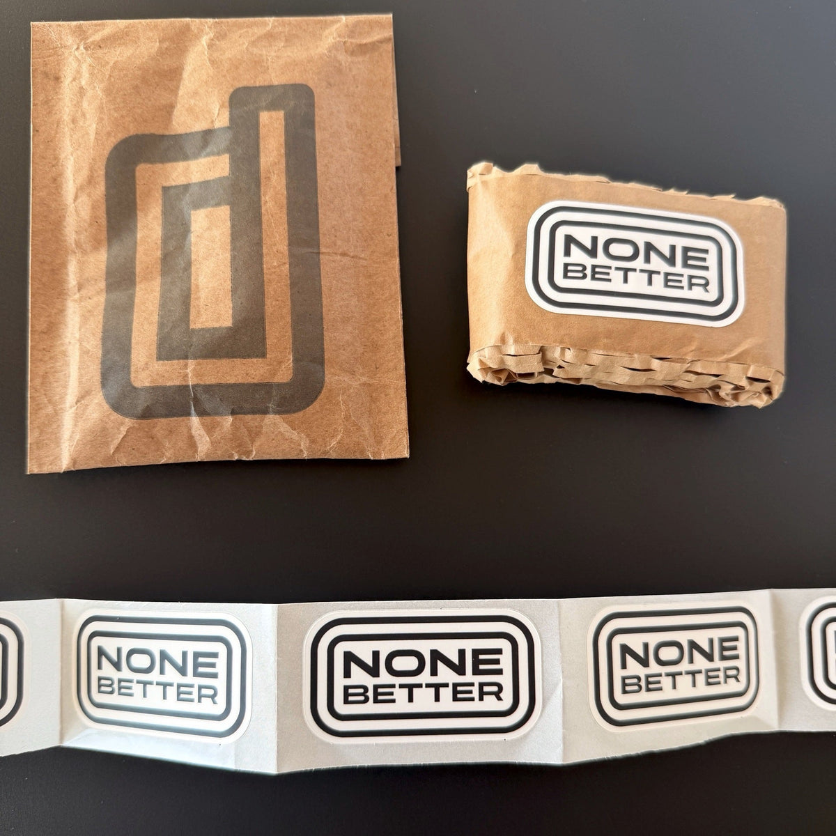 A kraft paper pouch, a honeycomb-wrapped bundle sealed with a None Better sticker, and a strip of stickers on a black board